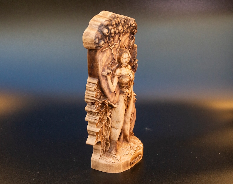 May include: A wooden sculpture of a woman standing in front of a tree. The sculpture is carved in detail and has a natural wood finish.