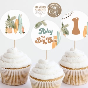May include: Three cupcakes topped with white frosting, each adorned with a circular topper. The toppers feature surfboards, palm fronds, and the text "Riley is a Big One" in a retro font. One topper displays the number "1".