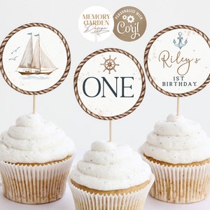 May include: Three cupcakes topped with nautical-themed decorations. The toppers feature a sailboat, a ship's wheel with the word "ONE", and an anchor with "Riley's 1st Birthday". Each topper has a rope-like border and is on a wooden stick.