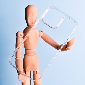 May include: A clear, transparent phone case held by a wooden mannequin. The case is rectangular with rounded corners and a camera cutout. The background is a light blue.