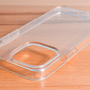 May include: A clear, transparent phone case made of rigid plastic. The case has a rectangular shape with rounded corners and a camera cutout. The case is designed to protect a smartphone and is shown on a wooden surface.
