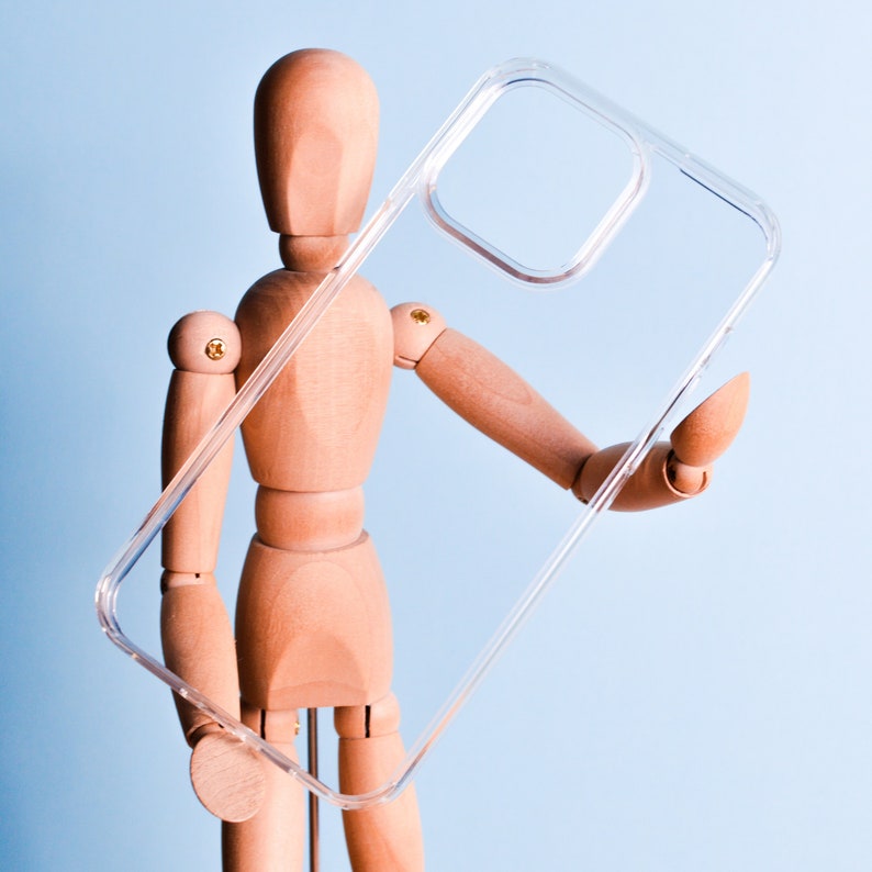 May include: A clear, transparent phone case held by a wooden mannequin. The case is rectangular with rounded corners and a camera cutout. The background is a light blue.