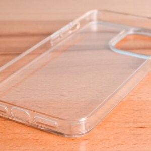May include: A clear, transparent phone case made of hard plastic. The case has cutouts for the camera and buttons. The phone case is shown on a light brown wooden surface.