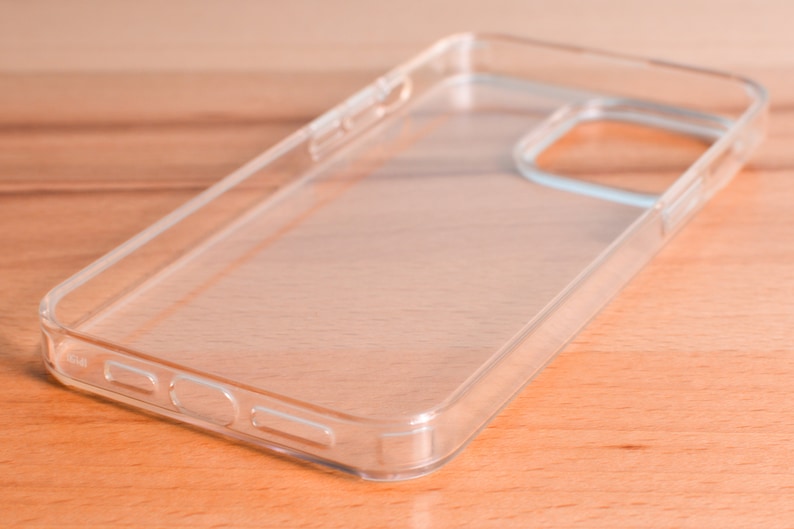 May include: A clear, transparent phone case made of hard plastic. The case has cutouts for the camera and buttons. The phone case is shown on a light brown wooden surface.