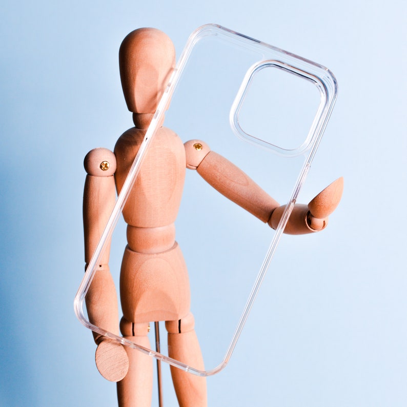 May include: A clear, transparent phone case held by a wooden mannequin. The case has a rectangular camera cutout and is designed to fit a smartphone. The background is a light blue.