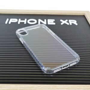 May include: Clear iPhone XR phone case. The case is transparent and made of a hard plastic material. The words "IPHONE XR" are displayed in white block letters on a black background.