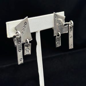 May include: A pair of silver earrings with a geometric design. Each earring features a small star charm and a dangling rectangular charm with the word "OFF" or "OOO" engraved on it.