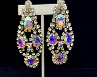 May include: A pair of dazzling chandelier earrings. Each earring features a teardrop design with iridescent, multi-colored stones and clear, sparkling crystals. The earrings are displayed on a white stand against a dark background.
