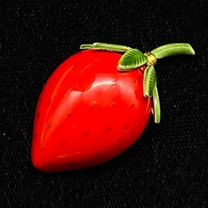 May include: A red and green ceramic strawberry brooch with a gold accent. The strawberry is detailed with small red seeds.