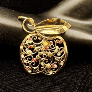 May include: A gold-toned apple-shaped brooch with intricate filigree detailing. The brooch features a leaf and stem design, adorned with clear and red gemstones. The overall design is ornate and vintage-inspired, suitable for adding a touch of elegance.