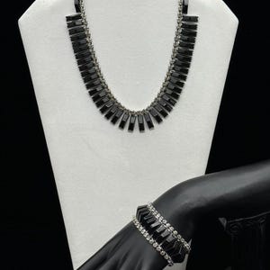 Könnte beinhalten: Ein schwarzes und silbernes Halsketten- und Armbandset mit geometrischem Design. Die Halskette hat ein fächerförmiges Design mit schwarzen Steinen und silbernen Akzenten. Das Armband ist eine doppelte Reihe schwarzer Steine mit silbernen Akzenten.