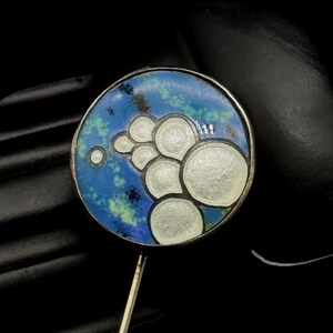 May include: A circular lapel pin featuring a blue and green mottled enamel background with a cluster of raised, off-white circular shapes. The pin has a thin metal stem extending from the bottom and a metallic border surrounding the circular face.