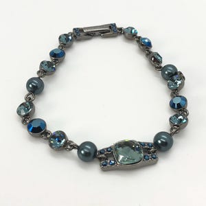 Puede incluir: Un brazalete gris oscuro con cuentas de cristal azul y turquesa y un cristal turquesa rectangular más grande en el centro.