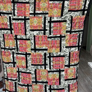 May include: A colourful quilt with a patchwork design. The quilt features squares of vibrant pink, yellow, and orange patterns, separated by black lines. The background is a grey and white pattern. The edges are trimmed with black lace.