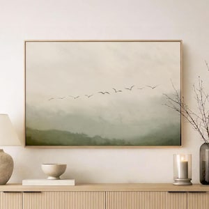 以下が含まれることがあります： 霧のかかった緑の山々の上を飛ぶ鳥の群れを描いた額入りの風景画。そのアートワークは、垂直の溝のあるドアが付いた明るい木製のキャビネットの上に掛けられています。キャビネットには、ランプ、ボウル、キャンドル、枝の入った花瓶があります。