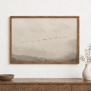 Peut inclure: Une impression murale rectangulaire dans un cadre en bois foncé représentant un paysage beige et taupe avec une volée d'oiseaux volant en ligne dans un ciel nuageux et brumeux. L'œuvre est exposée au-dessus d'une surface en bois avec un bol et un vase.