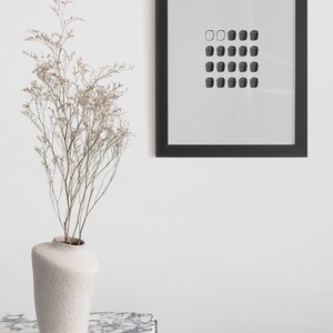 May include: A black framed print with a white background and a grid of 20 small black squares. The print is hanging on a white wall above a white marble table with a white ceramic vase with dried flowers.