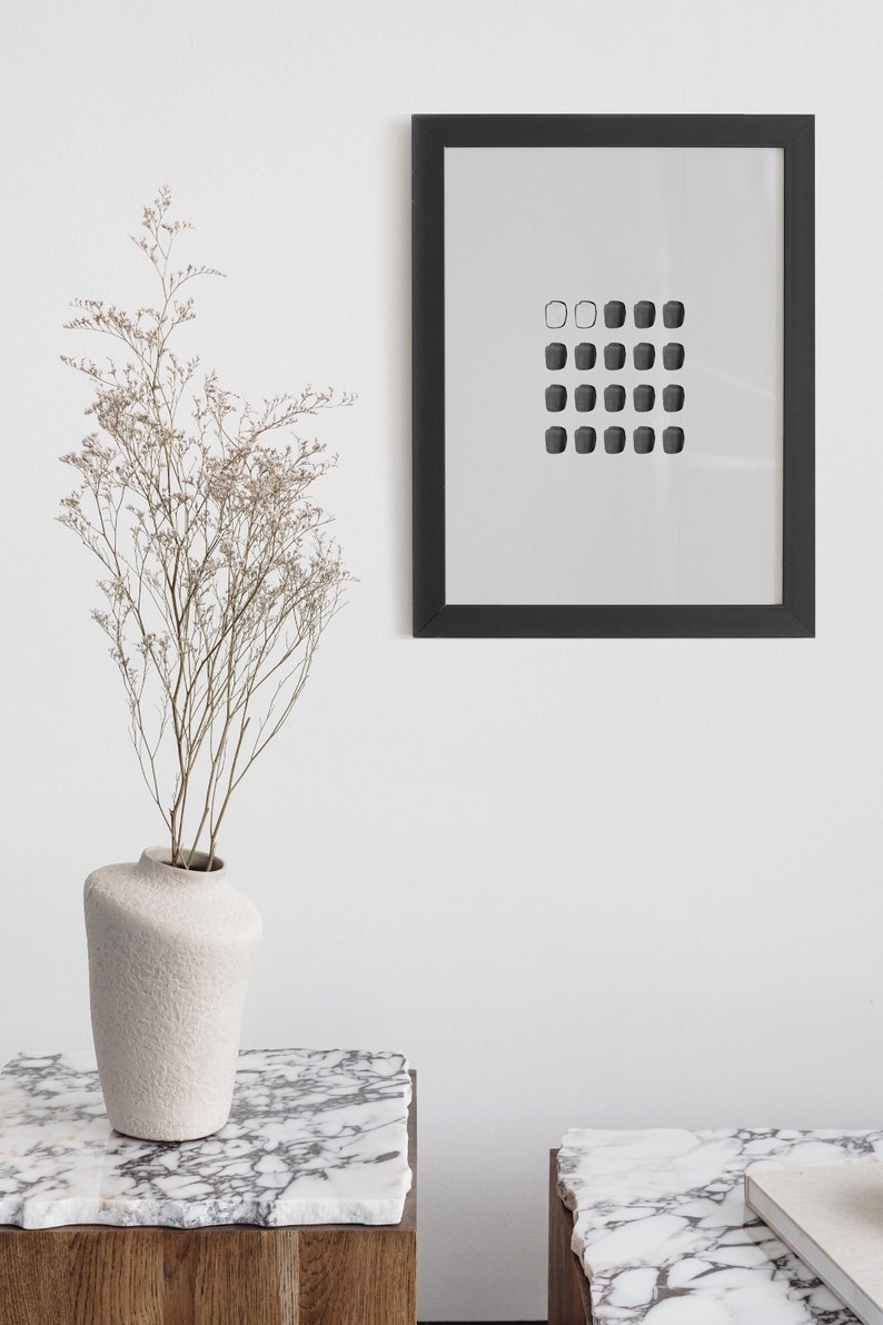 May include: A black framed print with a white background and a grid of 20 small black squares. The print is hanging on a white wall above a white marble table with a white ceramic vase with dried flowers.