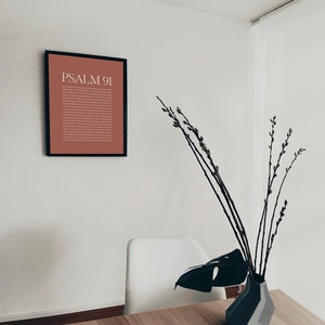 May include: A framed print with a brown background and white text that reads "PSALM 91". The print is hanging on a white wall. A white chair and a vase with branches and a large green leaf are on a wooden table.