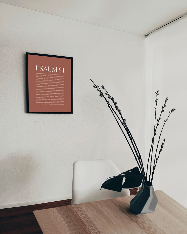 May include: A framed print with a brown background and white text that reads "PSALM 91". The print is hanging on a white wall. A white chair and a vase with branches and a large green leaf are on a wooden table.