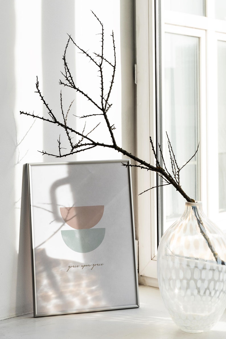 K&ouml;nnte beinhalten: Ein gerahmter Druck mit einem minimalistischen Design aus zwei sich &uuml;berlappenden Halbkreisen in Rosa- und Blaut&ouml;nen. Der Druck enth&auml;lt den Text "grace upon grace" in einer kursivschriftlichen Schriftart.