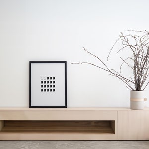 May include: A black framed picture with a white background and a grid of black and white squares hangs above a light wood shelf with a single open shelf below. A vase with a light brown band around it holds a dried branch with brown twigs.