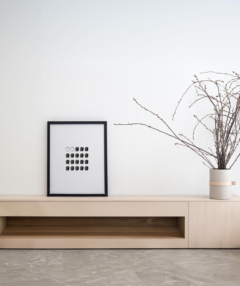 May include: A black framed picture with a white background and a grid of black and white squares hangs above a light wood shelf with a single open shelf below. A vase with a light brown band around it holds a dried branch with brown twigs.