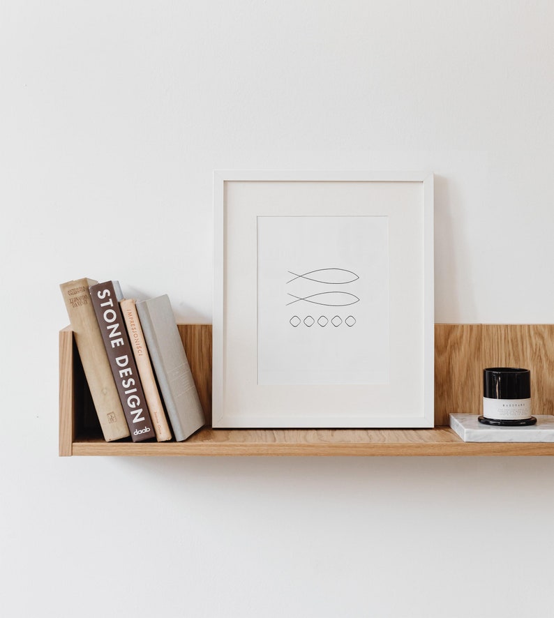 May include: A white framed print with a simple line drawing of three fish and five circles. The print is on a wooden shelf with a stack of books and a small black candle.