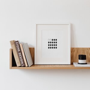 May include: A white framed print with a black and white geometric design of squares on a white background. The print is on a wooden shelf with a stack of books and a candle.