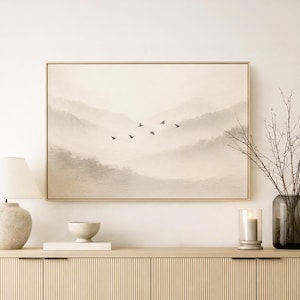 May include: A framed art print shows a tranquil mountain landscape in soft colours. A group of birds are seen in flight. The artwork is displayed above a light-coloured wooden cabinet, with a lamp and candle holder.