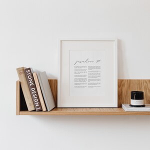 May include: A white framed print with the text "Psalm 91" in black cursive script. The print is on a wooden shelf with a stack of books and a black candle on a white marble surface.