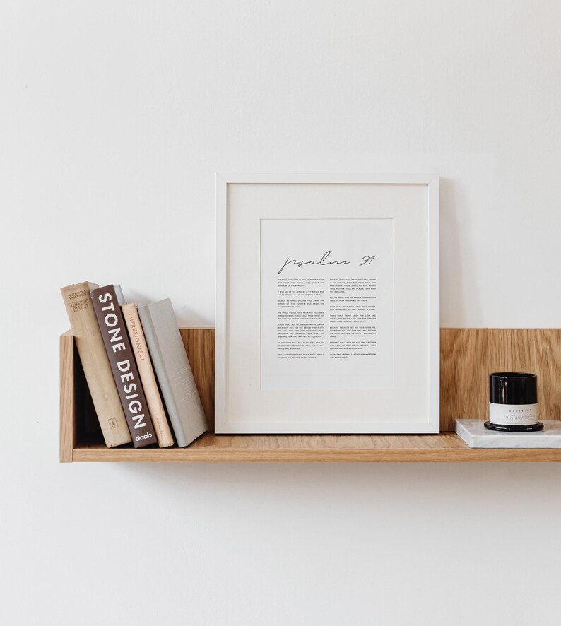 May include: A white framed print with the text "Psalm 91" in black cursive script. The print is on a wooden shelf with a stack of books and a black candle on a white marble surface.