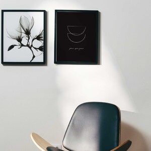 Puede incluir: Dos impresiones en blanco y negro enmarcadas en una pared blanca. La impresi&oacute;n de la izquierda presenta una imagen en blanco y negro de una flor de magnolia. La impresi&oacute;n de la derecha presenta un fondo negro con un dise&ntilde;o geom&eacute;trico blanco y el texto "grace upon grace". Una silla de cuero negro con patas de madera se encuentra frente a la pared.