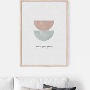K&ouml;nnte beinhalten: Ein gerahmter Druck mit einem minimalistischen Design, das zwei sich &uuml;berlappende Halbkreise in gedeckten Rosa- und Blaut&ouml;nen zeigt. Der Text "grace upon grace" ist in einer kursivschriftlichen Schriftart unter den Formen geschrieben.