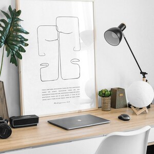 May include: A framed black and white line art print of two faces with a quote from Philippians 4:8. The print is on a white background and is framed in a light wood frame. The print is sitting on a desk with a laptop, a lamp, and a potted plant.
