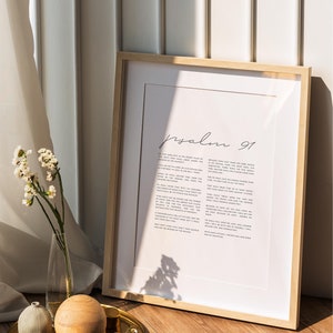 May include: A framed print of Psalm 91 in a light wood frame. The print is on white paper with black text. The frame is sitting on a wooden surface with a gold tray and decorative objects.
