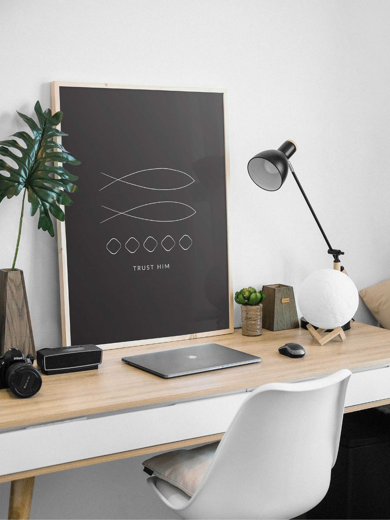 May include: A black and white minimalist poster with a line drawing of two fish and the text "TRUST HIM". The poster is framed in wood and is hanging on a white wall above a desk with a laptop, a lamp, and a potted plant.