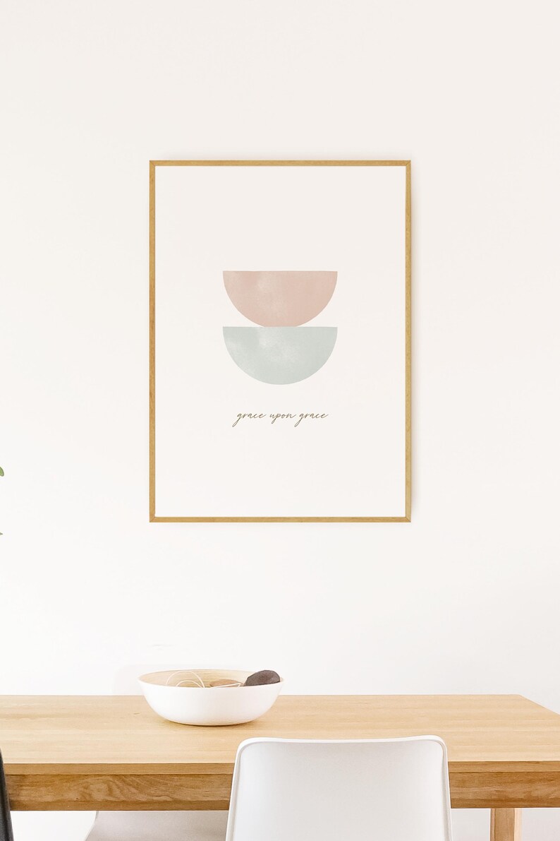 K&ouml;nnte beinhalten: Ein gerahmter Druck mit einem minimalistischen Design, das zwei sich &uuml;berlappende Halbkreise in Rosa- und Blaut&ouml;nen zeigt. Der Text "grace upon grace" ist in einer kursivschriftlichen Schriftart unter den Formen geschrieben.