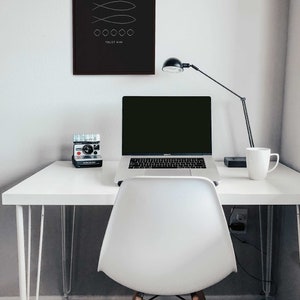 May include: A white desk with a laptop, a camera, a mug, and a lamp. The desk has a black frame and white legs. There is a white chair in front of the desk. A black framed print with the text "TRUST HIM" and a simple design of two fish is hanging on the wall behind the desk.