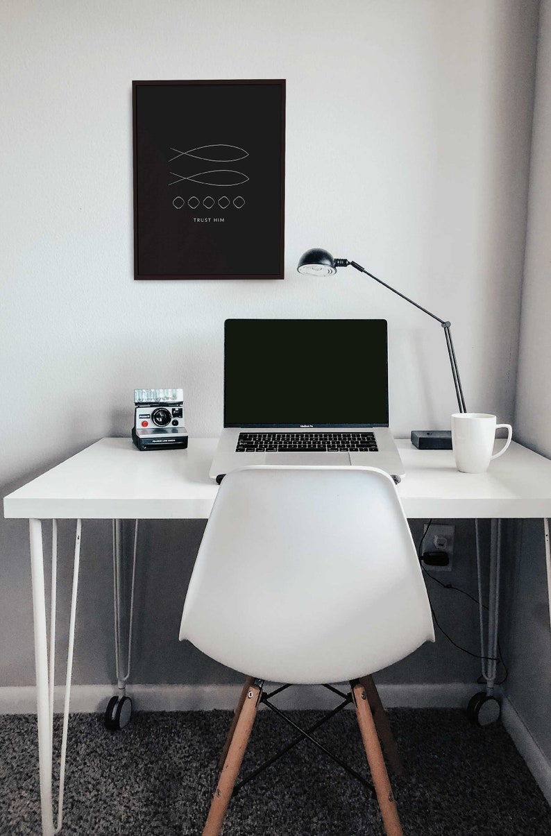 May include: A white desk with a laptop, a camera, a mug, and a lamp. The desk has a black frame and white legs. There is a white chair in front of the desk. A black framed print with the text "TRUST HIM" and a simple design of two fish is hanging on the wall behind the desk.