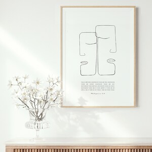May include: A framed print with a minimalist line drawing of two faces, with a quote from Philippians 4:8. The print is on a white background and has a light wood frame. The print is sitting on a wooden cabinet with a slatted front.