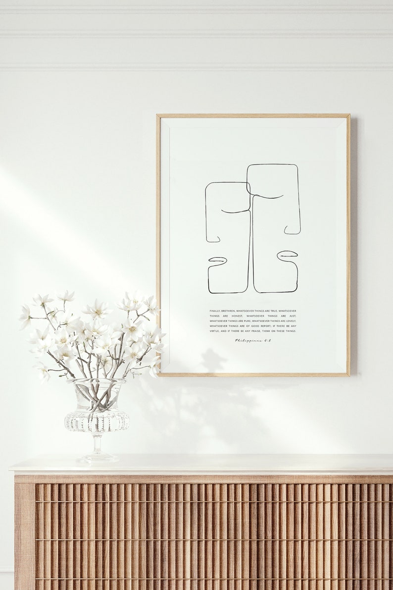May include: A framed print with a minimalist line drawing of two faces, with a quote from Philippians 4:8. The print is on a white background and has a light wood frame. The print is sitting on a wooden cabinet with a slatted front.