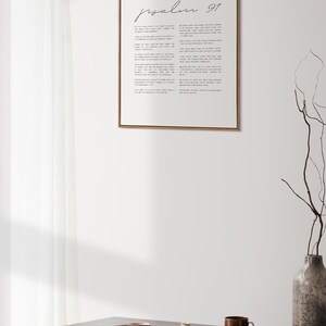 May include: A framed print of Psalm 91 in a minimalist style with black text on a white background. The print is hanging on a white wall above a wooden table with a vase, plates, and a cup.