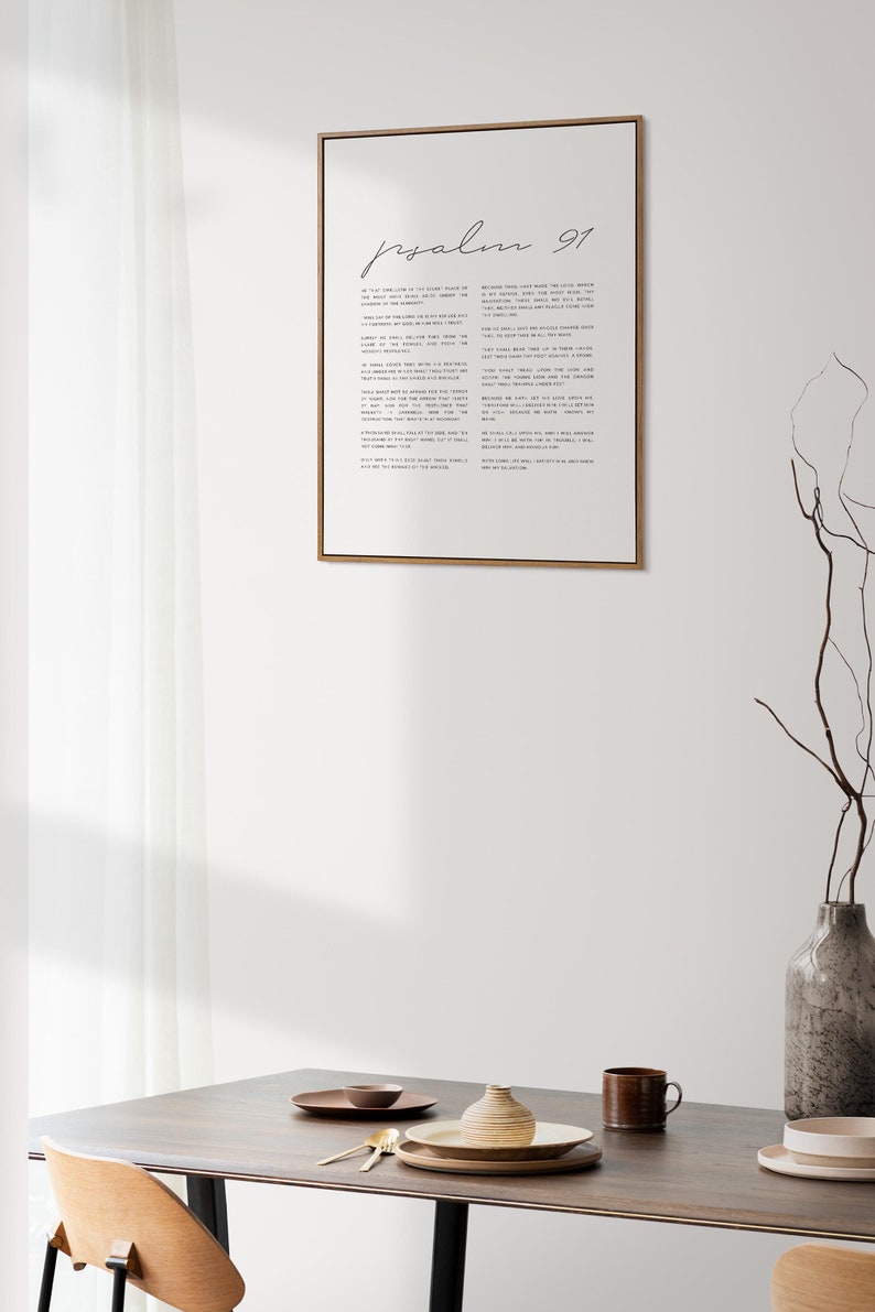 May include: A framed print of Psalm 91 in a minimalist style with black text on a white background. The print is hanging on a white wall above a wooden table with a vase, plates, and a cup.