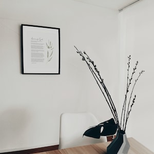 K&ouml;nnte beinhalten: Ein gerahmter Druck mit dem Text "Amor of God" und einer Aquarellillustration von gr&uuml;nen Bl&auml;ttern. Ein wei&szlig;er Stuhl und eine schwarze Vase mit einem Monstera-Blatt und Weidenzweigen stehen auf einem Holztisch.