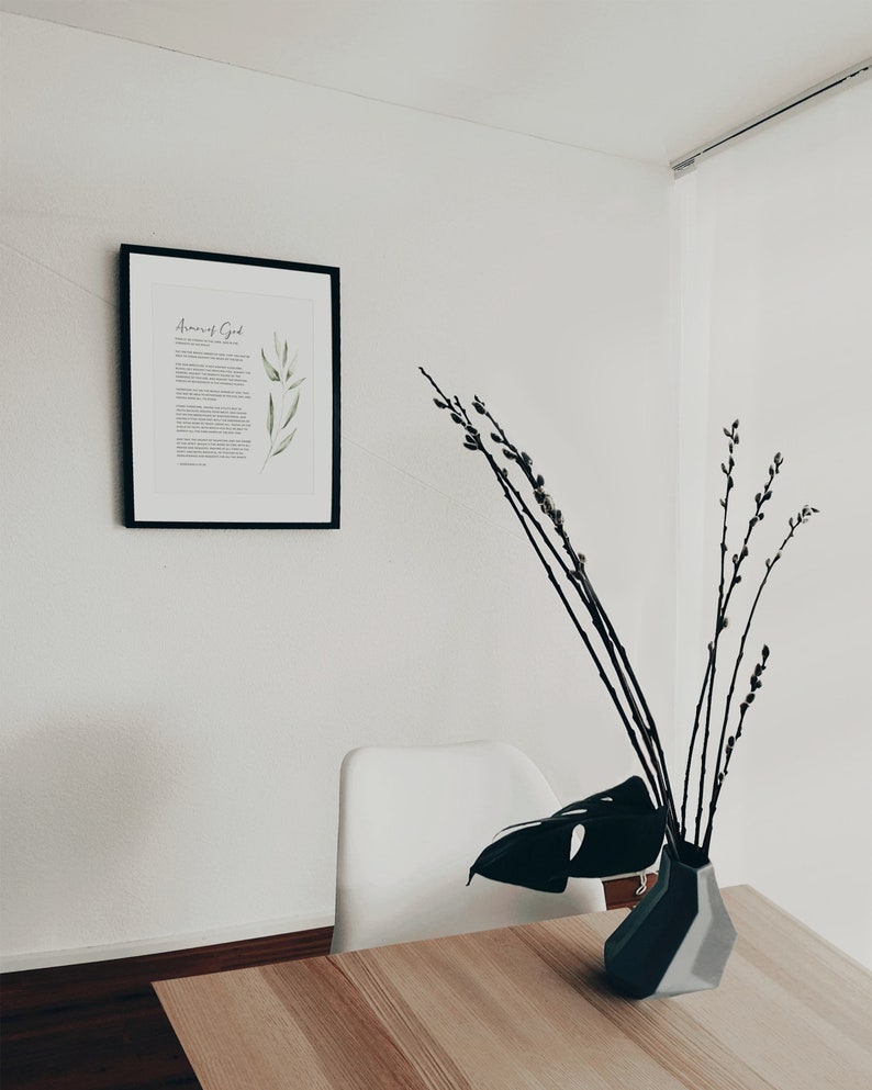 K&ouml;nnte beinhalten: Ein gerahmter Druck mit dem Text "Amor of God" und einer Aquarellillustration von gr&uuml;nen Bl&auml;ttern. Ein wei&szlig;er Stuhl und eine schwarze Vase mit einem Monstera-Blatt und Weidenzweigen stehen auf einem Holztisch.