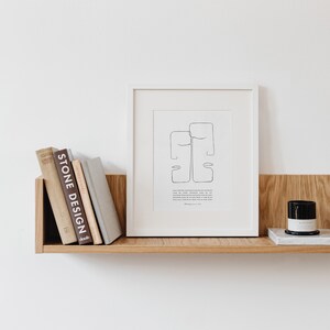 May include: A white framed print with a black and white line drawing of two faces. The print is on a wooden shelf with a candle and books.