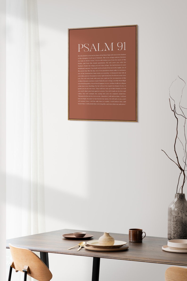 May include: A framed print with a brown background and white text. The text reads "PSALM 91" and includes a passage from the Bible. The print is hanging on a white wall above a wooden table with a vase and dishes.