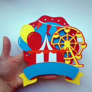 May include: A colorful paper craft featuring a red and white striped tent, a yellow Ferris wheel, and blue and yellow balloons. The craft has a blue banner with a blank space for text.