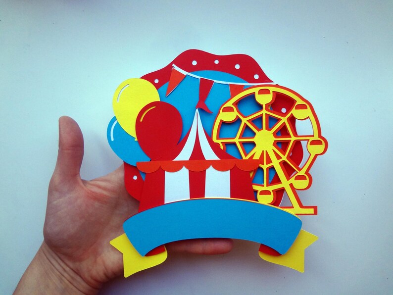 May include: A colorful paper craft featuring a red and white striped tent, a yellow Ferris wheel, and blue and yellow balloons. The craft has a blue banner with a blank space for text.